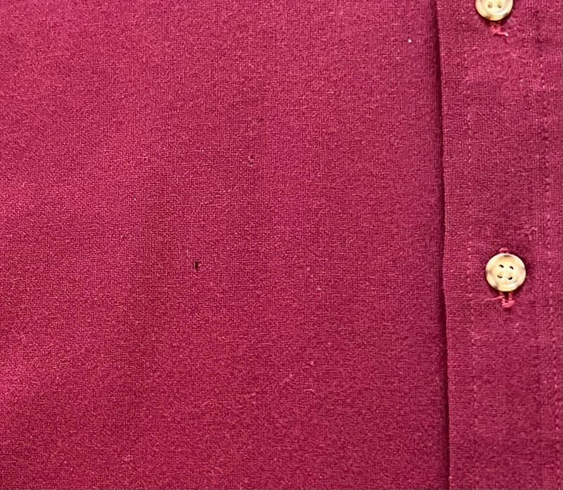 May include: A deep magenta or maroon colored shirt featuring a vertical placket with two visible light-colored, four-hole buttons. The fabric has a textured, slightly fuzzy appearance with a small, dark pinhole imperfection visible on the left side.
