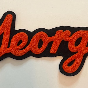May include: A black felt patch featuring the name George embroidered in bright orange chenille yarn. The lettering is in a cursive script style with a thick, textured pile, and the black backing follows the contour of the orange text.