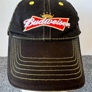 May include: Black baseball cap with yellow stitching and a curved bill. The cap features the Budweiser logo in white and red, with a gold crown above the brand name. The cap has a yellow button on top.