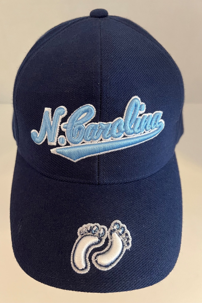 Pu&ograve; includere: Un cappello da baseball blu navy con un logo ricamato "N. Carolina" blu chiaro e un design di impronta di piede di bambino bianco sul davanti.