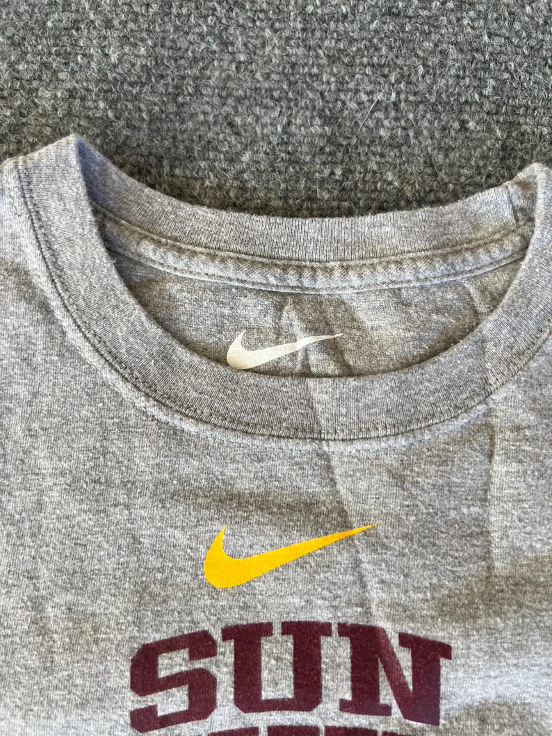 Pu&ograve; includere: Una maglietta grigia girocollo con il logo Nike bianco sul girocollo e un logo giallo con la scritta "SUN" in bordeaux. La maglietta &egrave; realizzata in un materiale morbido e confortevole.