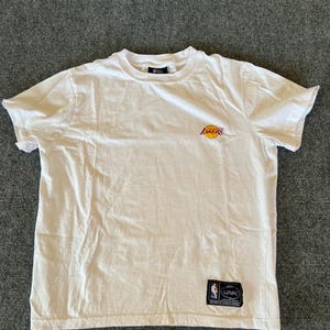 May include: White short-sleeved t-shirt with the Lakers logo in purple and gold on the chest. A small patch with the NBA logo is visible near the bottom. The shirt is made of a soft, lightweight material.