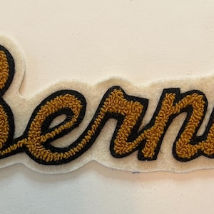 May include: A fabric patch featuring the name Bernie in a cursive script. The lettering is rendered in a textured gold thread with a thick black outline, all set against a cream-colored felt backing that follows the contour of the word.