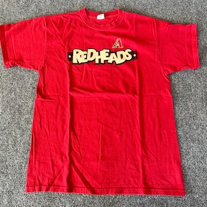 May include: A bright red t-shirt with the word "REDHEADS" in a black and yellow font across the chest. Above the word is a small logo. The shirt is made of a soft material and has short sleeves.