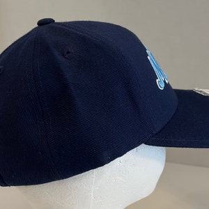 Pu&ograve; includere: Un cappello da baseball blu navy con un logo ricamato blu chiaro sul davanti. Il cappello ha una visiera curva e una cinghia regolabile.