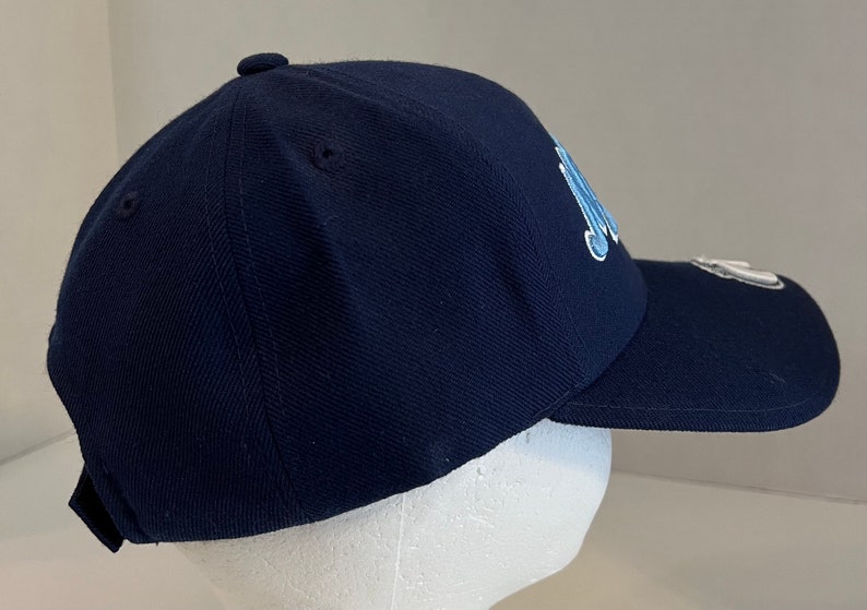 Pu&ograve; includere: Un cappello da baseball blu navy con un logo ricamato blu chiaro sul davanti. Il cappello ha una visiera curva e una cinghia regolabile.