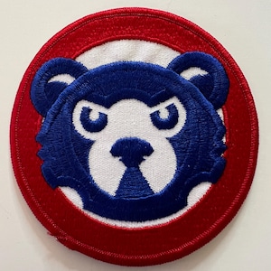 May include: A circular embroidered patch featuring a stylized blue bear head with white facial details. The bear is centered on a white background, which is surrounded by a thick red border. The entire design is finished with a red stitched edge.