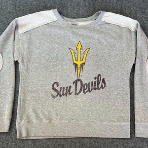 May include: Gray sweatshirt with white patches on the shoulders and elbows. The front features a yellow trident logo and the words "Sun Devils" in a distressed font. A crew neck sweatshirt.