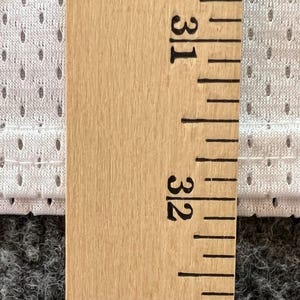 Op de afbeelding: Close-up van een houten liniaal met inch-metingen, naast een witte, geperforeerde stof. De stof heeft kleine, gelijkmatig verdeelde gaatjes. De liniaal geeft een meting van 31 tot 32 inch (78,7 tot 81,3 cm) aan.