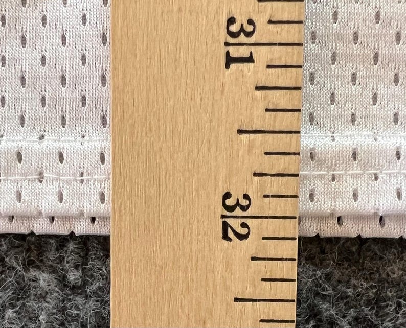 Op de afbeelding: Close-up van een houten liniaal met inch-metingen, naast een witte, geperforeerde stof. De stof heeft kleine, gelijkmatig verdeelde gaatjes. De liniaal geeft een meting van 31 tot 32 inch (78,7 tot 81,3 cm) aan.