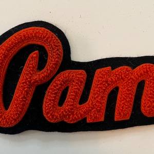 May include: A black fabric patch featuring the name Pam embroidered in bright orange, textured, raised yarn. The lettering is written in a cursive script style, and the black backing follows the contour of the orange embroidered name.