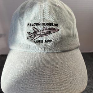 May include: A light blue baseball cap with the text "FALCON DUNES DC LUKE AFB" embroidered above a jet illustration. The cap has a curved brim and a fabric strap closure.