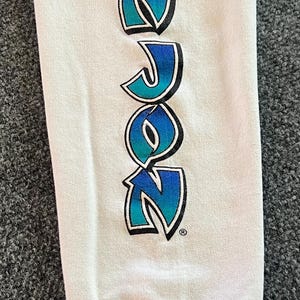 Peut inclure: Une manche de chemise &agrave; manches longues blanche avec le texte RON JON imprim&eacute; verticalement dans une police audacieuse et stylis&eacute;e. Les lettres passent d'un bleu sarcelle vif en haut &agrave; un bleu roi profond en bas, soulign&eacute;es de noir.