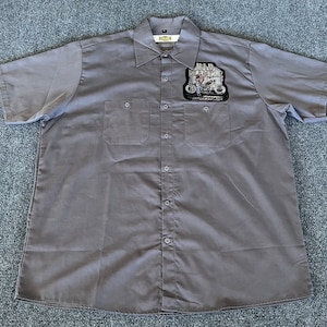 May include: Gray short-sleeve button-up shirt with two chest pockets. A patch on the left chest reads "BAD MONSTER" with a motorcycle graphic. The shirt has a classic collar and button closures.