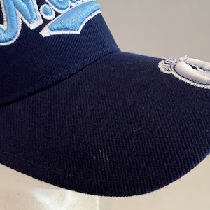 Pu&ograve; includere: Un cappello da baseball blu navy con un logo ricamato in azzurro e bianco sul davanti. Il logo dice "Nova" in un font stilizzato.