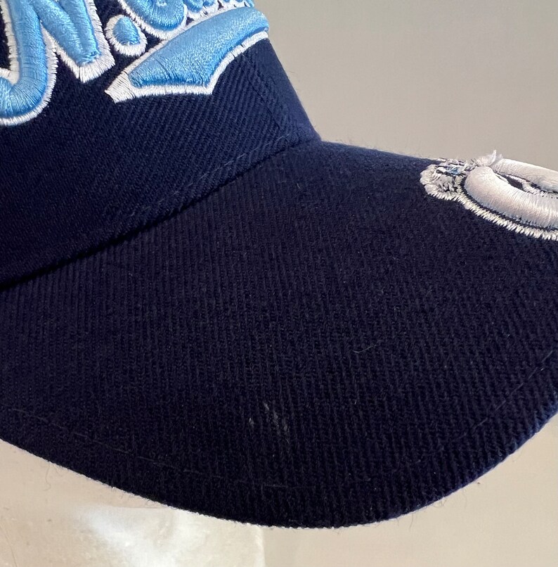 Pu&ograve; includere: Un cappello da baseball blu navy con un logo ricamato in azzurro e bianco sul davanti. Il logo dice "Nova" in un font stilizzato.