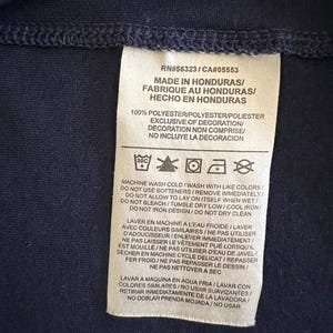 Op de afbeelding: Een marineblauw kledinglabel met tekst in het Engels, Frans en Spaans. Het label geeft aan dat het artikel in Honduras is gemaakt en 100% polyester is. Wasvoorschriften staan ook op het label.