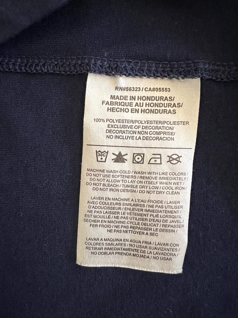 Op de afbeelding: Een marineblauw kledinglabel met tekst in het Engels, Frans en Spaans. Het label geeft aan dat het artikel in Honduras is gemaakt en 100% polyester is. Wasvoorschriften staan ook op het label.