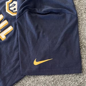 Op de afbeelding: Marineblauw T-shirt met een gouden Nike swoosh-logo op de mouw. De shirt heeft een grote, gedeeltelijk zichtbare afbeelding aan de linkerbovenkant met het woord "BALL" in goud.