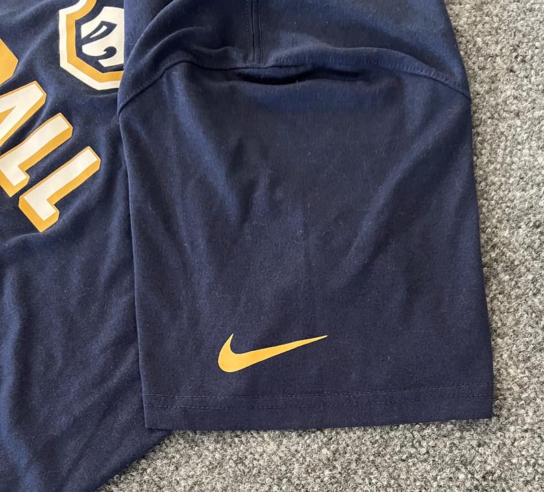 Op de afbeelding: Marineblauw T-shirt met een gouden Nike swoosh-logo op de mouw. De shirt heeft een grote, gedeeltelijk zichtbare afbeelding aan de linkerbovenkant met het woord "BALL" in goud.