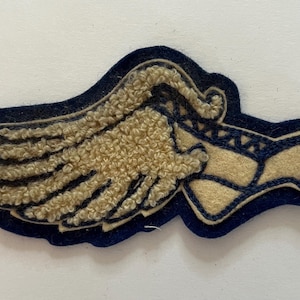 May include: A navy blue and beige felt patch featuring a stylized winged shoe design. The patch has a dark navy blue base layer with a textured beige felt wing and a beige shoe outline with stitched detailing, creating a layered appearance.