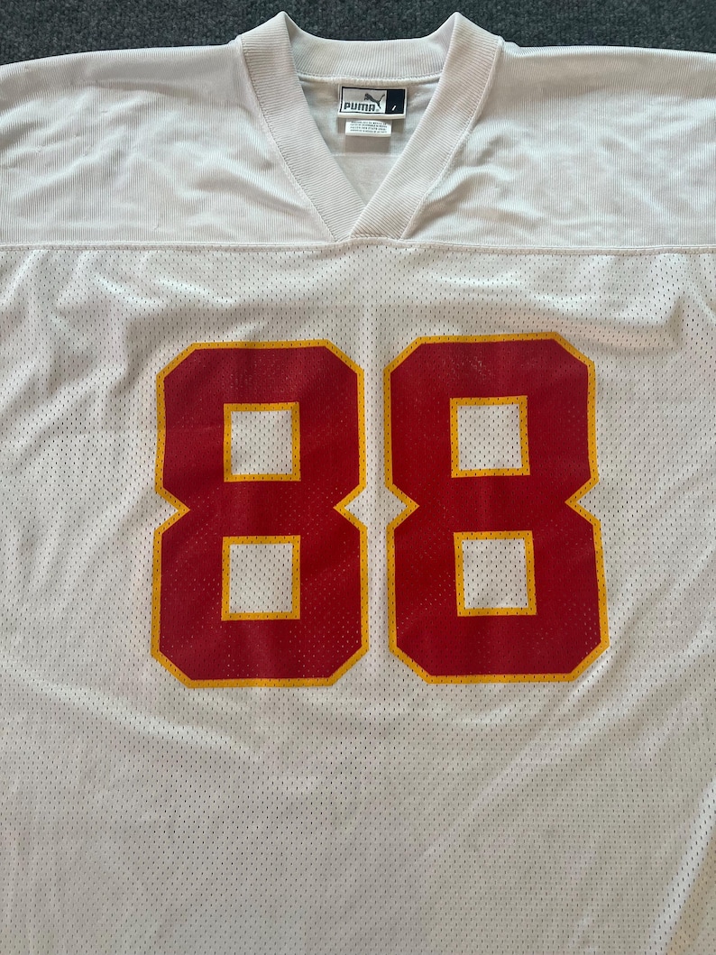 Op de afbeelding: Wit mesh sportshirt met het nummer 88 in rood met gele randen. Het shirt heeft een V-hals en een Puma-logo. De stof heeft een geperforeerde textuur.