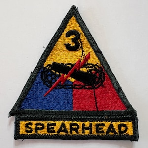 May include: A triangular embroidered patch featuring a yellow top section with a black number 3, a blue lower left section, and a red lower right section. A black tank silhouette and a red lightning bolt are centered. A yellow tab reads SPEARHEAD.