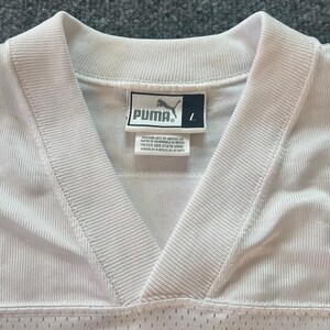Op de afbeelding: Close-up van een wit Puma-shirt met een V-hals. Het Puma-logo is zichtbaar op een label, samen met de maat "L". Het shirt heeft een geribbelde textuur en een mesh onderkant.