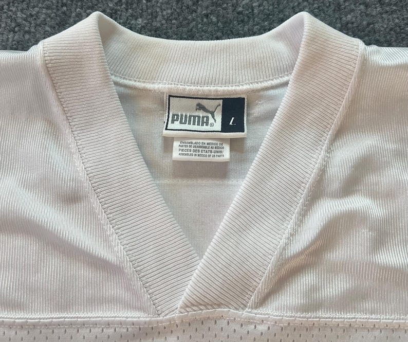 Op de afbeelding: Close-up van een wit Puma-shirt met een V-hals. Het Puma-logo is zichtbaar op een label, samen met de maat "L". Het shirt heeft een geribbelde textuur en een mesh onderkant.