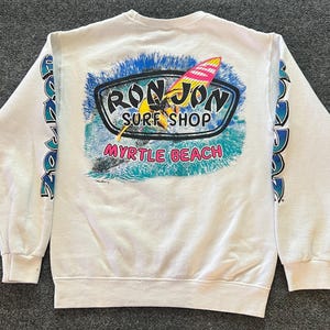 Peut inclure: Un sweat-shirt blanc &agrave; manches longues avec un motif au dos portant l'inscription Ron Jon Surf Shop et Myrtle Beach. Le motif repr&eacute;sente une personne faisant de la planche &agrave; voile sur des vagues bleues. Le texte Ron Jon est imprim&eacute; sur les manches.