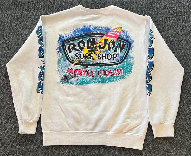 Peut inclure: Un sweat-shirt blanc &agrave; manches longues avec un motif au dos portant l'inscription Ron Jon Surf Shop et Myrtle Beach. Le motif repr&eacute;sente une personne faisant de la planche &agrave; voile sur des vagues bleues. Le texte Ron Jon est imprim&eacute; sur les manches.