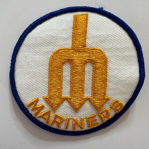 May include: A circular embroidered patch featuring a white background with a blue stitched border. In the center, a gold trident emblem is displayed above the word MARINERS, which is also stitched in gold thread in a curved arrangement.