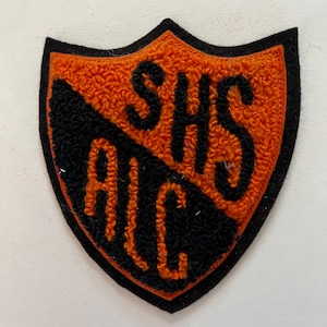 May include: A shield-shaped patch featuring a textured orange and black design. The upper portion displays the letters SHS in black, while the lower diagonal section features the letters ALC in orange, all set against a contrasting background.