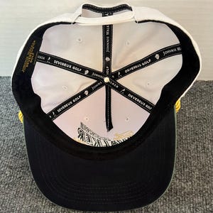 May include: A white and black baseball cap with the text "JOHNNIE WALKER" and "DEVEREUX GOLF" printed on the inside. The cap has a black brim and a white top. The cap is designed for golf.