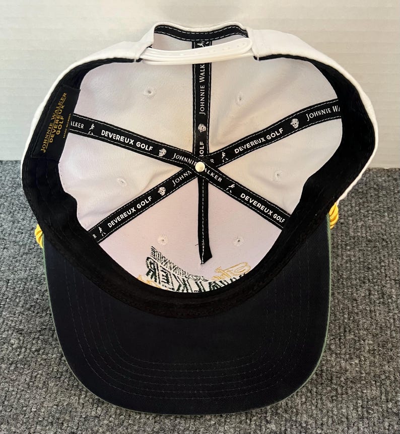 May include: A white and black baseball cap with the text "JOHNNIE WALKER" and "DEVEREUX GOLF" printed on the inside. The cap has a black brim and a white top. The cap is designed for golf.