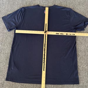 Op de afbeelding: Een marineblauw T-shirt met korte mouwen en een ronde hals. Een houten meetinstrument ligt over het shirt. Het shirt heeft een klein logo op de linkerbovenborst. Het shirt ligt op een grijze ondergrond.