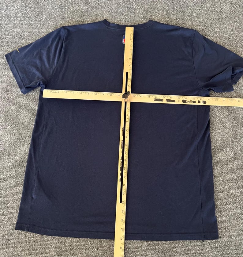 Op de afbeelding: Een marineblauw T-shirt met korte mouwen en een ronde hals. Een houten meetinstrument ligt over het shirt. Het shirt heeft een klein logo op de linkerbovenborst. Het shirt ligt op een grijze ondergrond.