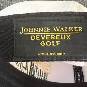 May include: Black baseball cap with the text "JOHNNIE WALKER DEVEREUX GOLF" in gold lettering on a black patch. The cap is white on the inside and has a tag that says "MADE IN CHINA".