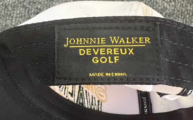 May include: Black baseball cap with the text "JOHNNIE WALKER DEVEREUX GOLF" in gold lettering on a black patch. The cap is white on the inside and has a tag that says "MADE IN CHINA".