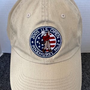 May include: Tan baseball cap with a curved brim and adjustable strap. The cap features an embroidered patch with the text "2005 U.S. OPEN PINEHURST No.2" and a golfer graphic in red, white, and blue.