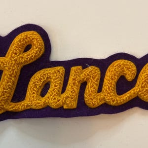 May include: A custom embroidered patch featuring the name Lance in a textured, golden-yellow cursive script. The lettering is layered onto a dark purple felt backing that follows the contour of the text, creating a bold, raised appearance.