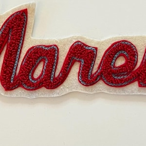 May include: A red and white embroidered patch featuring the cursive text "Mareno" in red thread with a light blue outline, set against a white felt backing. A wooden ruler is positioned vertically next to the patch, showing a length of 4 inches.