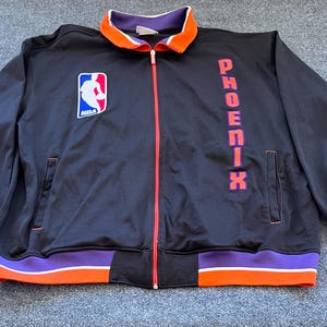 May include: Black zip-up jacket with orange and purple accents. Features the NBA logo and the word "PHOENIX" in orange lettering down the right side. The collar is orange and white.