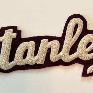 May include: A decorative patch featuring the name Stanley in cream-colored, textured, raised chenille lettering. The script text is outlined by a dark maroon felt backing that follows the contour of the letters, creating a bold contrast.