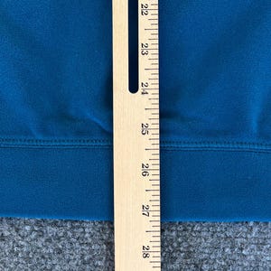 May include: A close-up of a teal blue fabric with a seam running horizontally across the center. A wooden ruler with markings from 22 to 28 is positioned vertically on the right side of the image.