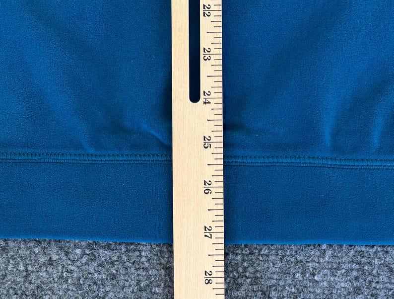 May include: A close-up of a teal blue fabric with a seam running horizontally across the center. A wooden ruler with markings from 22 to 28 is positioned vertically on the right side of the image.