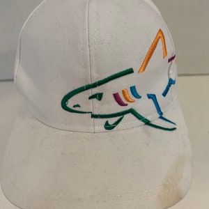 May include: White baseball cap with a colorful embroidered shark design. The shark is outlined in green, blue, orange, and purple.