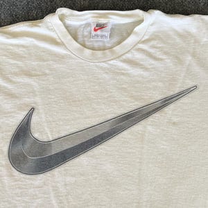 May include: Cream-colored t-shirt featuring a large, gradient gray Nike swoosh logo. The shirt has a crew neck and short sleeves. The Nike logo is a prominent design element, adding a sporty aesthetic to the casual wear.