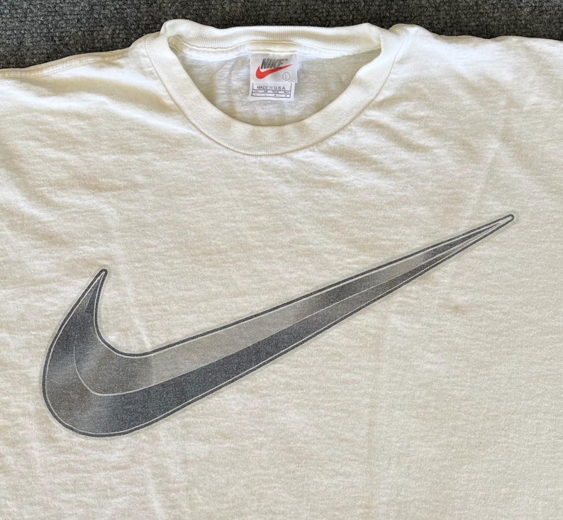May include: Cream-colored t-shirt featuring a large, gradient gray Nike swoosh logo. The shirt has a crew neck and short sleeves. The Nike logo is a prominent design element, adding a sporty aesthetic to the casual wear.