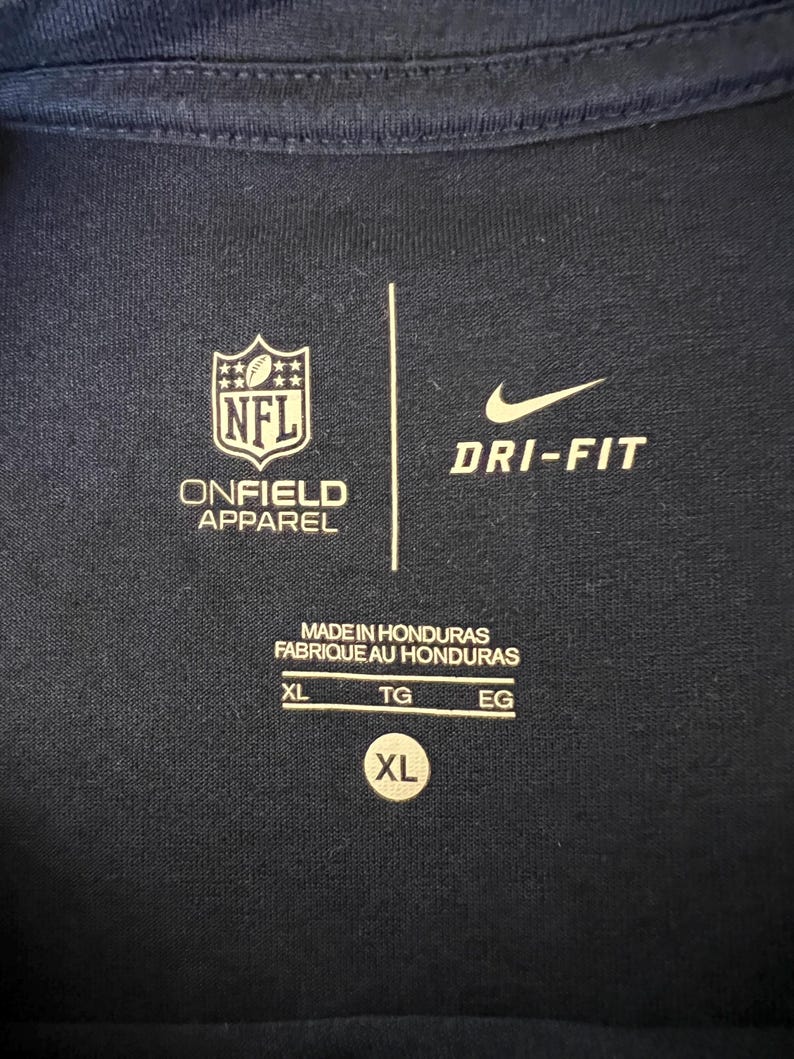 Op de afbeelding: Marineblauw sportshirt met het NFL-logo en het Nike Dri-FIT-logo. Het shirt heeft de tekst "ONFIELD APPAREL" en "MADE IN HONDURAS". De maat is XL.